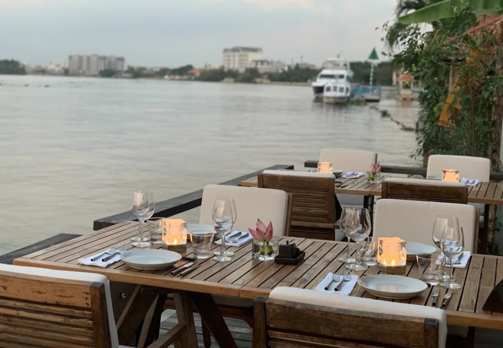 Một bức ảnh chụp không gian ngoài trời của nhà hàng The Deck Saigon, với bàn ghế gỗ, khăn trải bàn trắng, và đèn lồng trang trí. Nền ảnh là cảnh sông Sài Gòn và thành phố.