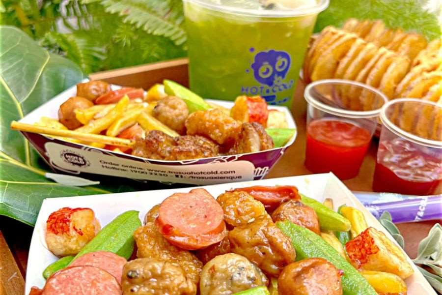 Một đĩa thức ăn đa dạng với nhiều loại hải sản, xúc xích, thịt viên, rau củ và khoai tây chiên, được bày biện trên một chiếc đĩa đen. Bên cạnh là một cốc nước ép xanh và một ly nước chấm.
