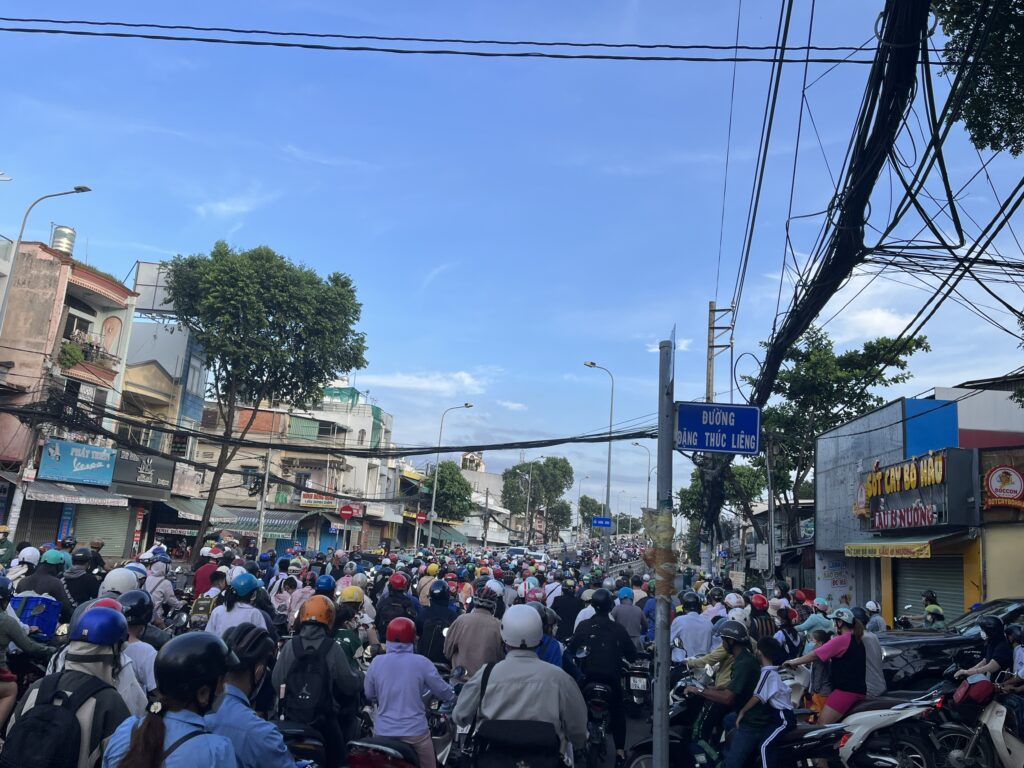 Cảnh đường phố đông đúc tại Việt Nam với tình trạng kẹt xe dưới chân cầu Chánh Hưng, phương tiện chủ yếu gồm các xe máy. Đường phố được bao quanh bởi các tòa nhà cũ, trong đó có các cửa hàng nhỏ. Biển tên đường nổi bật ghi "Đường Đặng Thúc Liêng."