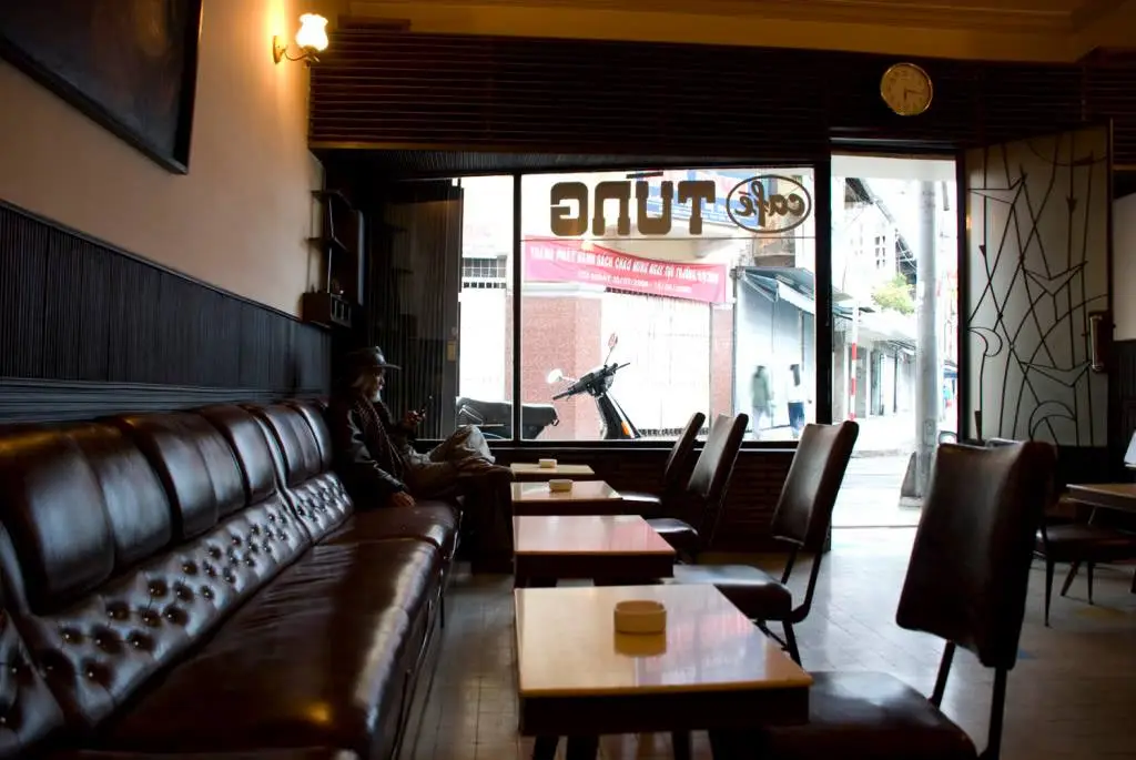 Không gian bên trong quán cafe Đà Lạt với nội thất cổ điển, ghế da màu nâu và ánh sáng trầm lắng, nhìn ra con phố yên tĩnh qua ô cửa kính lớn.