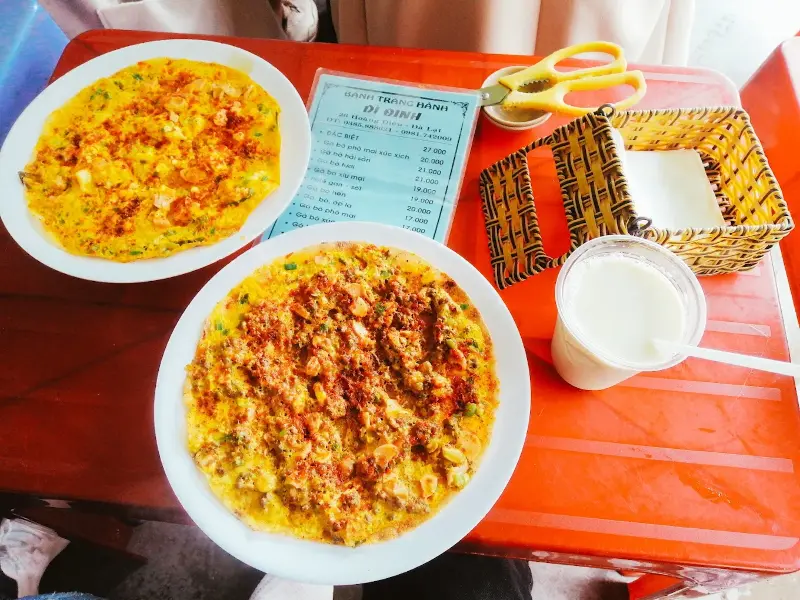 Top những quán ăn vặt Đà Lạt giá rẻ, ăn là ghiền Hai đĩa bánh tráng nướng cùng ly sữa đậu nành trên bàn nhựa đỏ tại một quán ăn vặt Đà Lạt nổi tiếng.