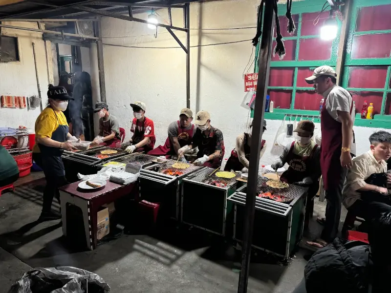 Quán bánh tráng nướng đông khách đang hoạt động nhộn nhịp vào buổi tối, nổi bật trong không gian ăn vặt Đà Lạt.