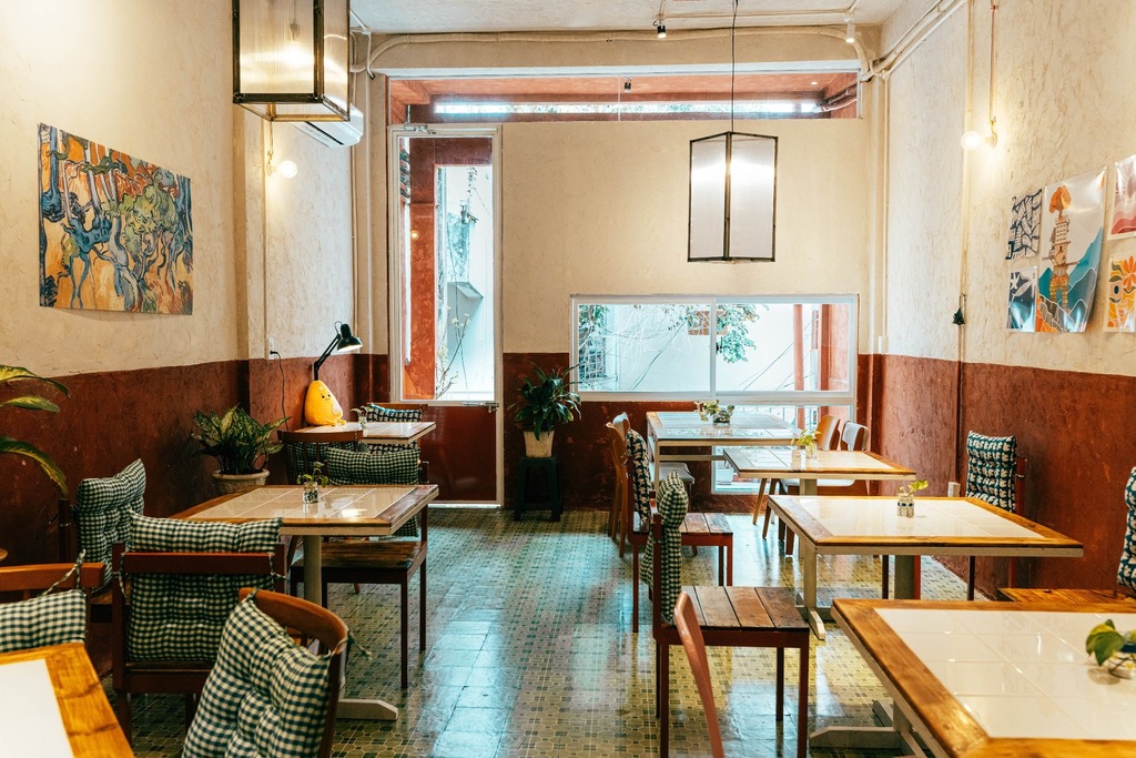 Không gian quán cafe với bàn gỗ, tranh treo tường nghệ thuật và ánh sáng tự nhiên ấm áp