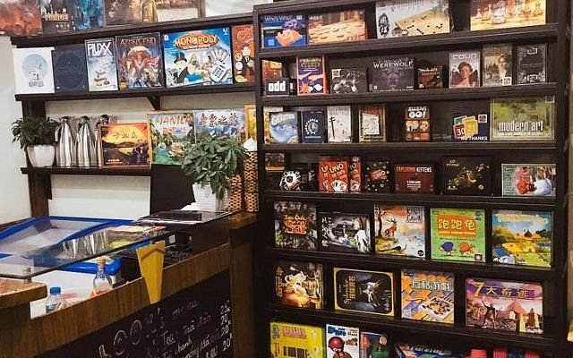 Không gian bên trong Board Game Station với các kệ gỗ cao chất đầy các hộp trò chơi board game đa dạng, bàn ghế gỗ xếp gọn gàng và ánh đèn vàng tạo cảm giác ấm cúng.
