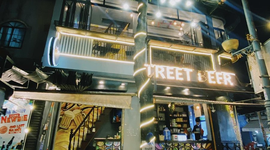 Không gian quán Street Beer ngoài trời với bàn ghế gỗ thấp và nhiều loại vỏ chai bia trang trí trên kệ.