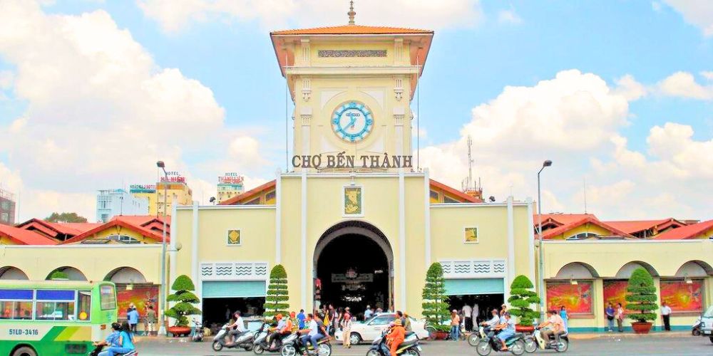 Cửa Nam chợ Bến Thành với tháp đồng hồ biểu tượng, khung cảnh giao thông nhộn nhịp phía trước và bầu trời xanh trong vắt.