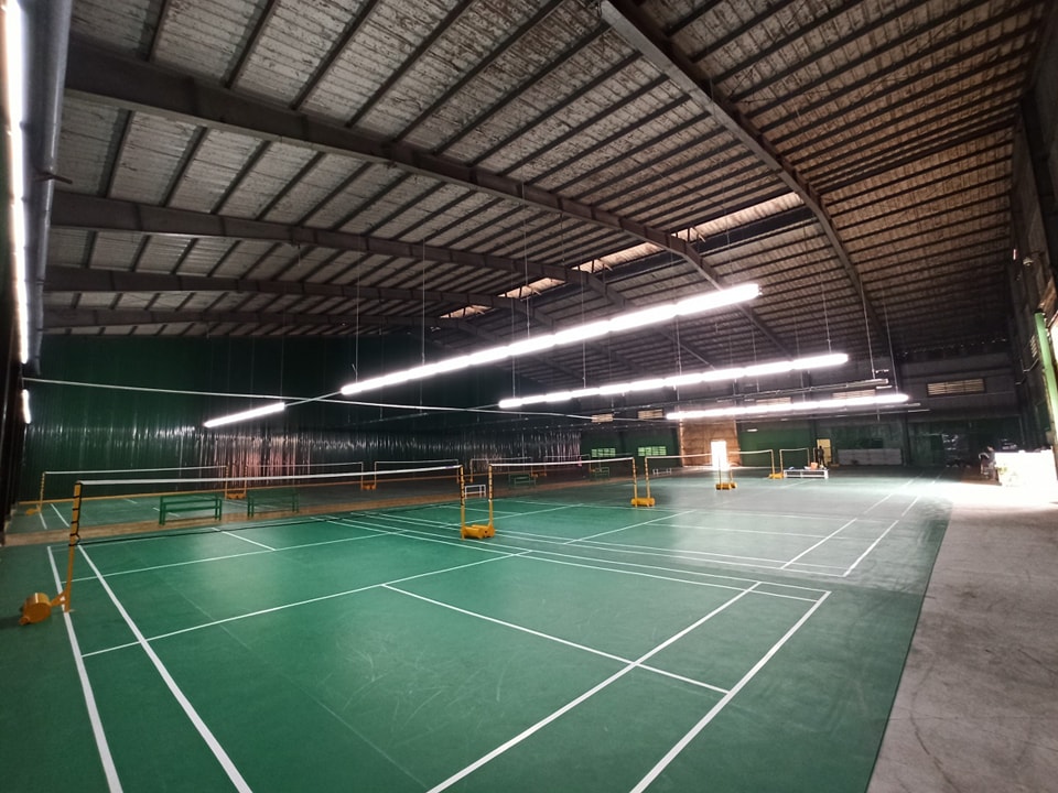 Các thảm sân cầu lông màu xanh lá tiêu chuẩn trong nhà với hệ thống lưới ngăn, vạch kẻ sơn trắng và đèn chiếu sáng phía trên cao.