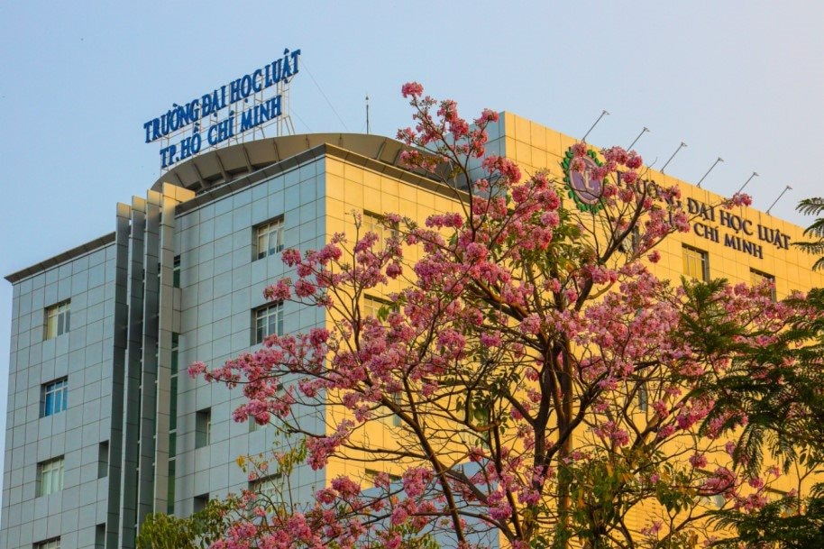 Tòa nhà Trường Đại học Luật TP.HCM với cây hoa nở phía trước và biển tên trên mái