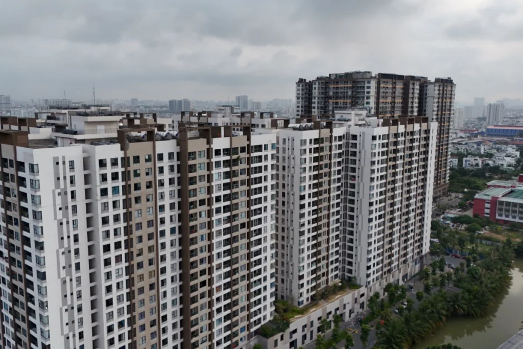 Tổ hợp các block chung cư cao tầng san sát nhau với ban công kính hiện đại, phía góc phải có một nhánh sông nhỏ uốn lượn và hàng cây xanh chạy dọc bờ kè dưới bầu trời mây xám