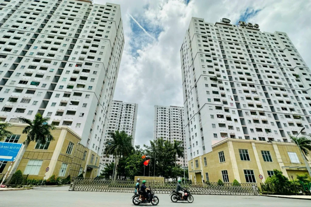 Hai tòa nhà chung cư cao tầng màu trắng xám xây sát nhau, phía dưới có cổng sắt xếp và hai người đi xe máy trên đường lộ nội khu