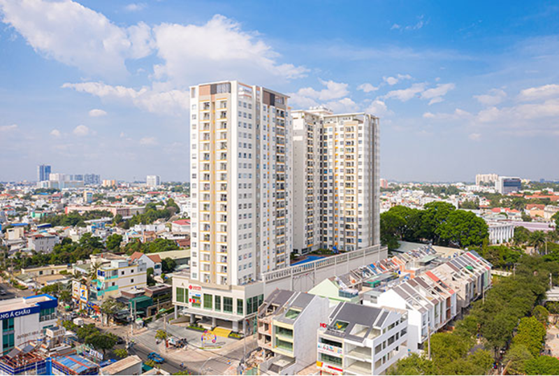 Toàn cảnh thực tế tòa nhà chung cư Moonlight Residences Thủ Đức cao tầng màu trắng xám dưới nền trời xanh có dãy nhà phố thương mại liền kề phía trước