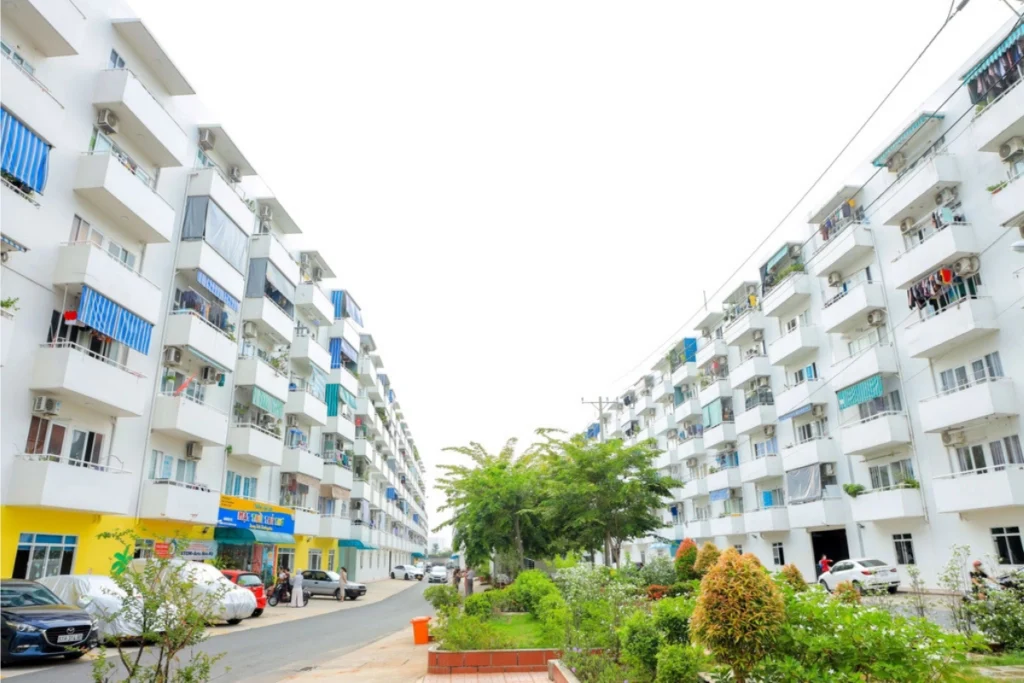 Góc nhìn từ giữa con đường nội khu hướng lên bầu trời trắng sáng, hai bên là hai dãy block chung cư thấp tầng sơn màu trắng đối diện nhau với các ban công nhỏ nhô ra