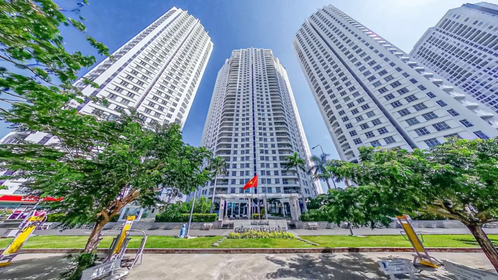 Hình ảnh thực tế từ góc thấp nhìn lên ba tòa tháp căn hộ Hoàng Anh Thanh Bình Quận 7 sơn màu trắng sừng sững dưới bầu trời xanh, phía trước là bãi cỏ và hàng cây xanh mát.