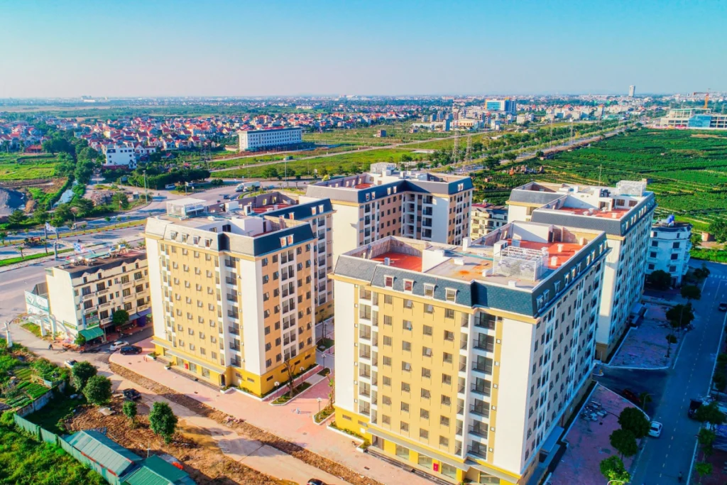 Góc nhìn trên cao xuống quần thể các block chung cư cao tầng màu vàng kem nhạt kết hợp mái ngói xám đen, bao quanh bởi hệ thống đường sá nội khu và ruộng đồng xanh mướt phía xa