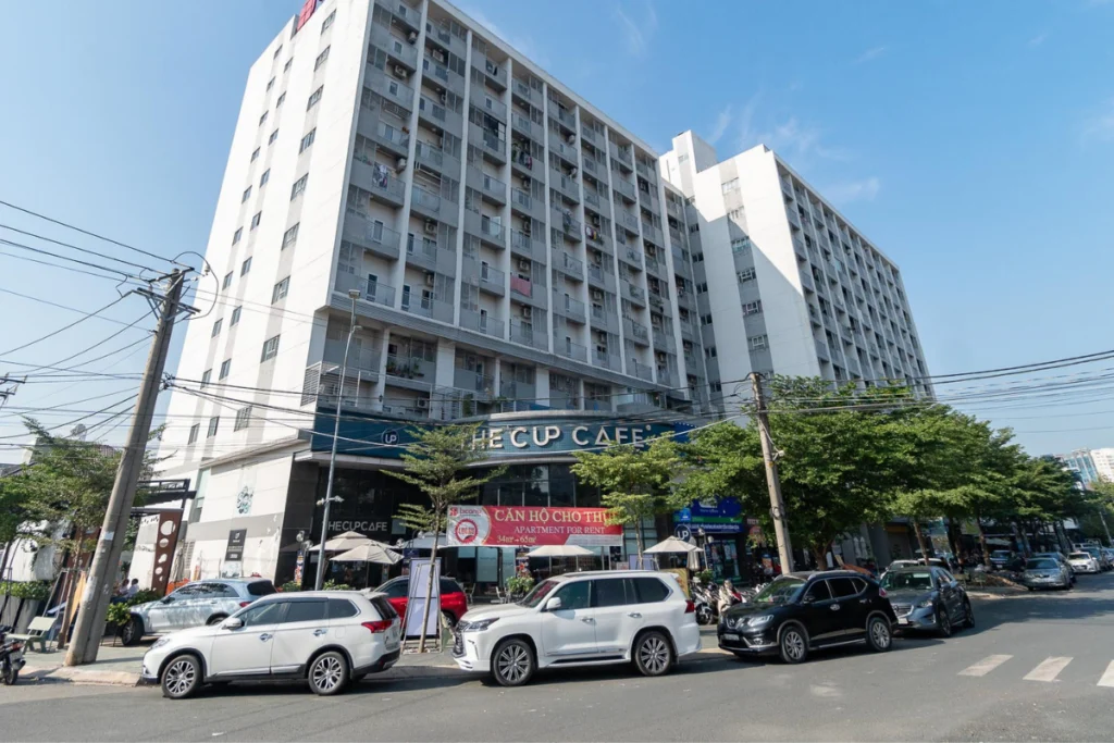 Ảnh chụp thực tế từ góc đường đối diện nhìn lên khối tòa nhà căn hộ Phú Hòa Lợi, phía dưới chân đế là quán cà phê sầm uất với dù che ngoài trời