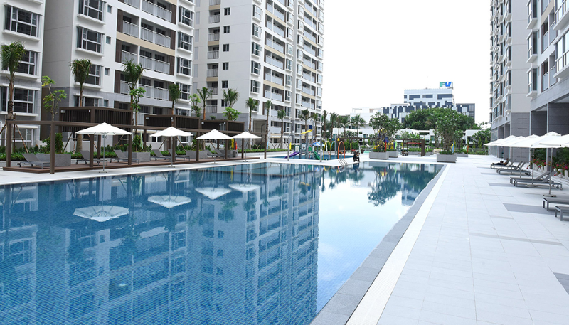 Hình ảnh thực tế hồ bơi ngoài trời xanh biếc tại chung cư Scenic Valley Quận 7 với hàng ghế tắm nắng, ô che nắng màu trắng và các block căn hộ bao quanh