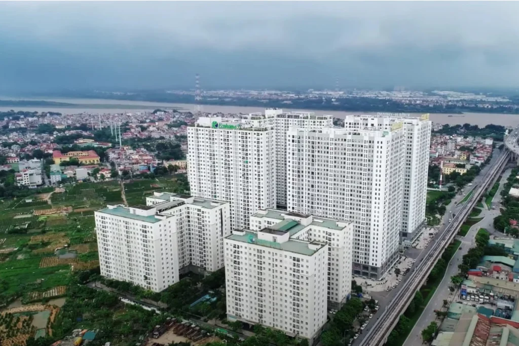 oàn cảnh cụm chung cư cao tầng hiện đại sơn màu trắng xám, nằm sát trục đường giao thông có tuyến đường sắt trên cao, phía xa là dòng sông lớn bao quanh khu dân cư