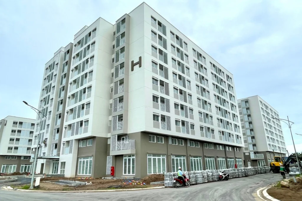 Dãy tòa nhà chung cư nhà ở xã hội cao khoảng 7 tầng với tông màu trắng chủ đạo pha xám ở chân đế, , phía dưới đường nội khu có pallet vật liệu xây dựng xếp ngay ngắn