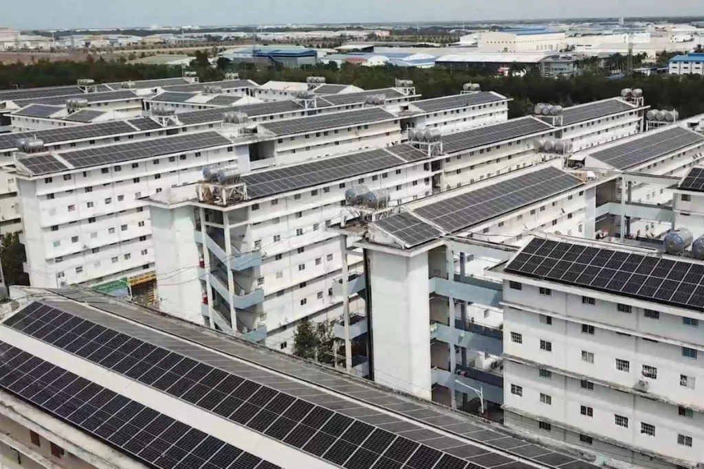 Góc nhìn từ trên cao xuống các block nhà chung cư thấp tầng sơn màu trắng có mái lợp các tấm pin năng lượng mặt trời xếp san sát nhau, phía xa là khu nhà xưởng công nghiệp