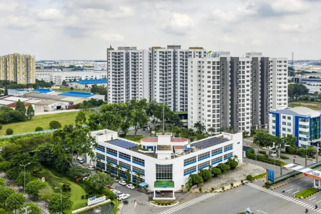 Góc nhìn từ trên cao xuống một tòa nhà chung cư cao tầng hiện đại tên Habitat đang được xây dựng bên cạnh các khu nhà xưởng công nghiệp thấp tầng, xung quanh có mảng xanh rộng lớn và đường quốc lộ chạy qua