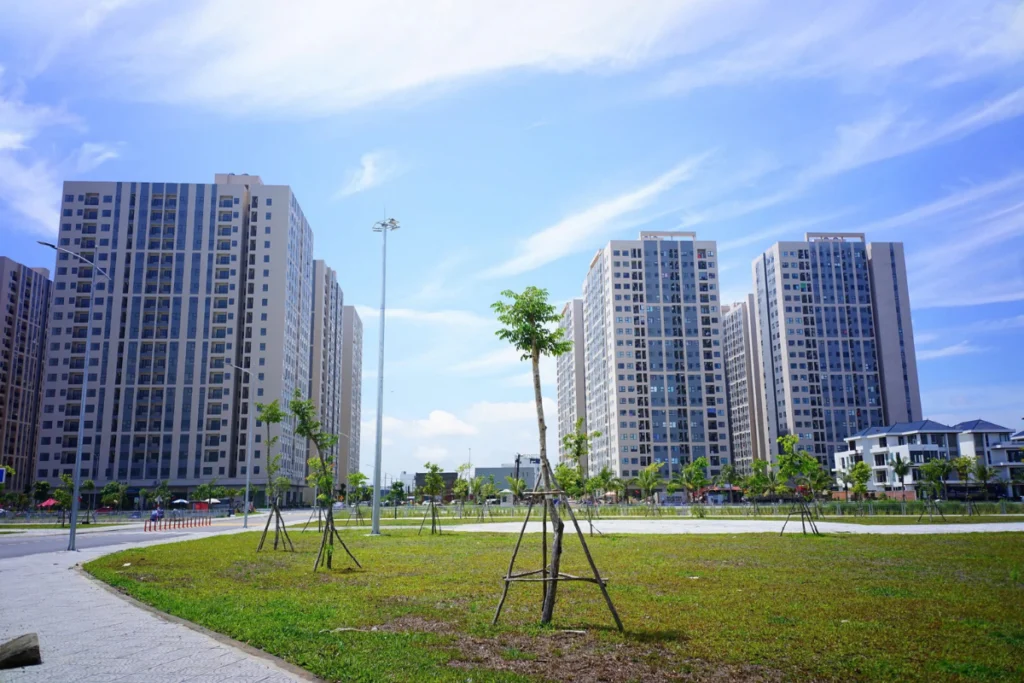 Khuôn viên quảng trường xanh mát với bãi cỏ rộng và hàng cây mới trồng phía trước dãy chung cư nhà ở xã hội cao tầng