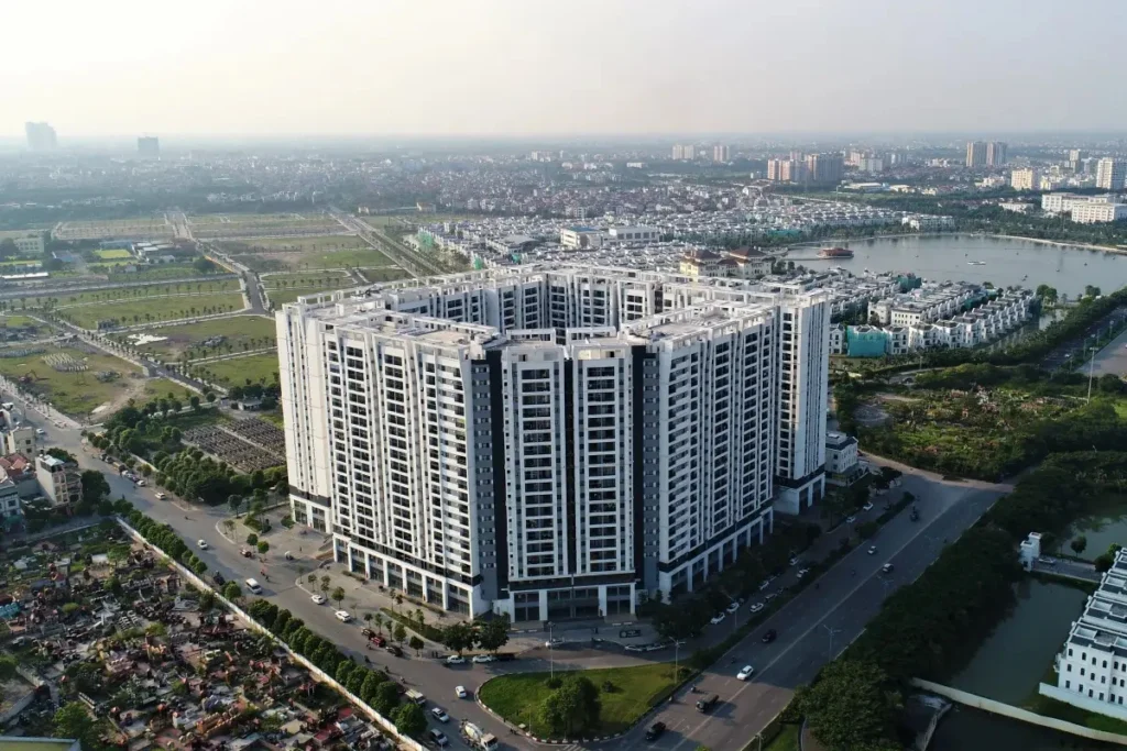 Khối tòa nhà chung cư lớn màu trắng xám được xây dựng vuông vức bao quanh một khoảng sân rộng ở giữa, tọa lạc sát tuyến đường lớn bên cạnh một hồ nước và khu đô thị thấp tầng