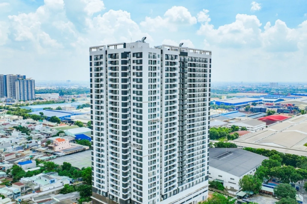 Góc chụp từ trên cao một tòa nhà chung cư cao tầng hiện đại với hai tông màu trắng và xám đen xen kẽ, xung quanh là khu dân cư và các nhà xưởng công nghiệp thấp tầng trải dài dưới bầu trời xanh