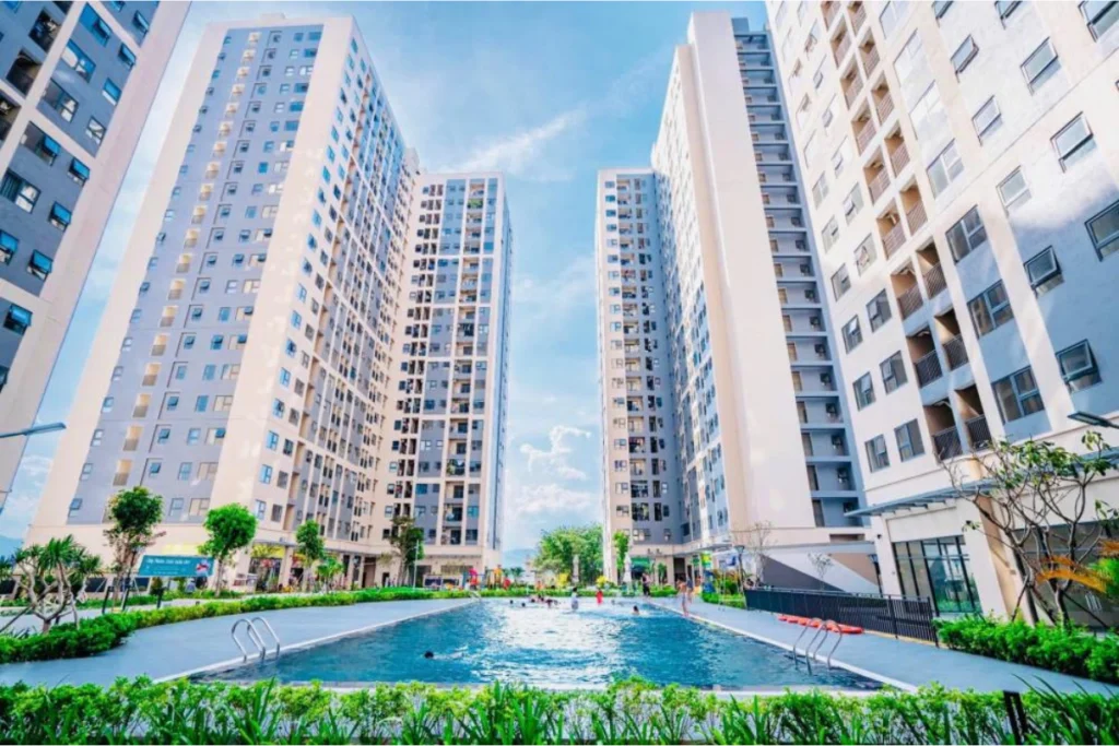 Ảnh chụp góc rộng từ hồ bơi nội khu nhìn lên các tòa tháp căn hộ chung cư cao tầng hiện đại với tông màu trắng chủ đạo đan xen các mảng sơn xám trung tính