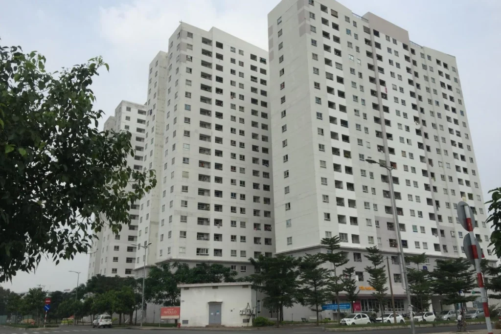 Ảnh chụp thực tế góc rộng từ bên kia đường nhìn sang dãy các tòa tháp chung cư cao tầng màu trắng xám khối hộp đứng vững chãi