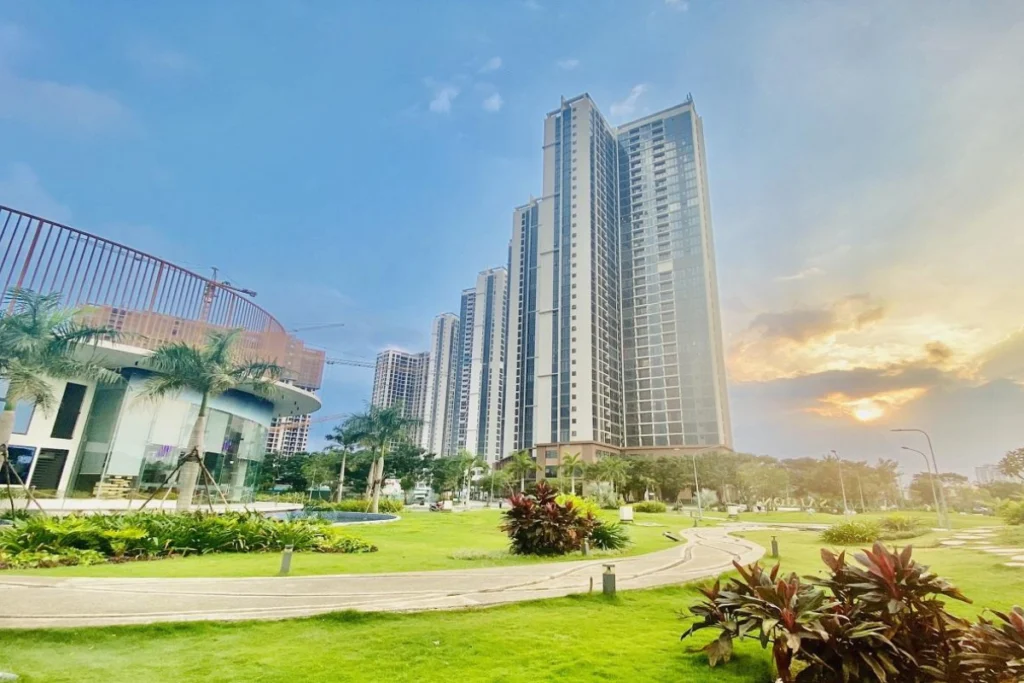 Ảnh thực tế khuôn viên công viên cây xanh nội khu rộng rãi với lối đi dạo uốn lượn, thảm cỏ xanh mướt, phía sau là các tòa tháp chung cư cao tầng hiện đại