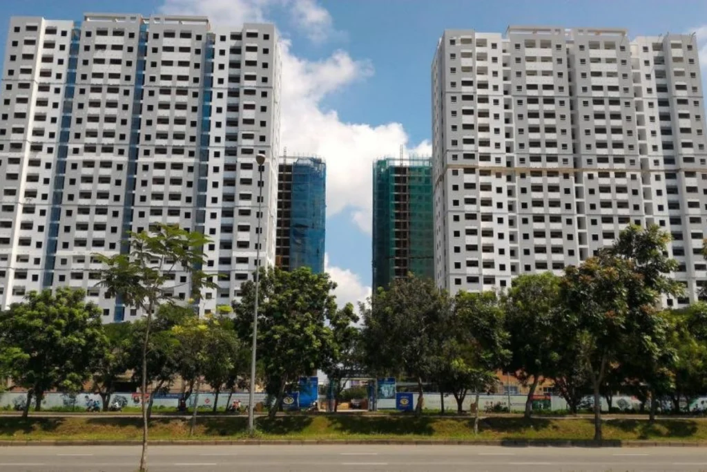 Toàn cảnh hai block chung cư cao tầng màu trắng xám đã hoàn thiện phần thô bên cạnh các block khác đang thi công, phía trước có hàng cây xanh dọc theo tuyến đường lộ