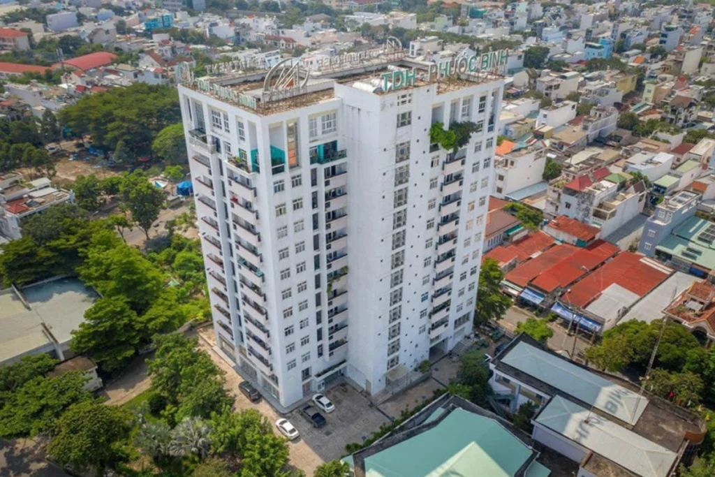 Toàn cảnh tòa nhà chung cư cao tầng TDH Phước Bình màu trắng nổi bật giữa khu dân cư, xung quanh có nhiều cây xanh và nhà thấp tầng, phía dưới sân có vài xe ô tô đang đỗ