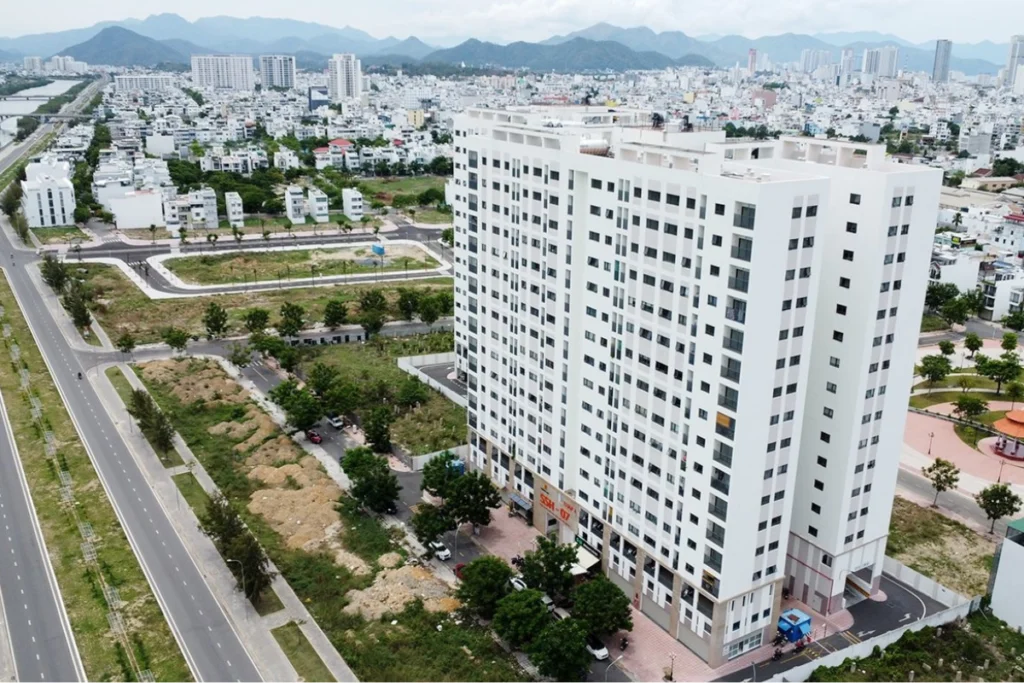 Góc nhìn từ trên cao xuống một tòa nhà chung cư cao tầng màu trắng hiện đại dáng chữ L, tọa lạc cạnh các tuyến đường giao thông rộng lớn, xung quanh có các bãi đất trống đang quy hoạch và đồi núi mờ ảo phía xa