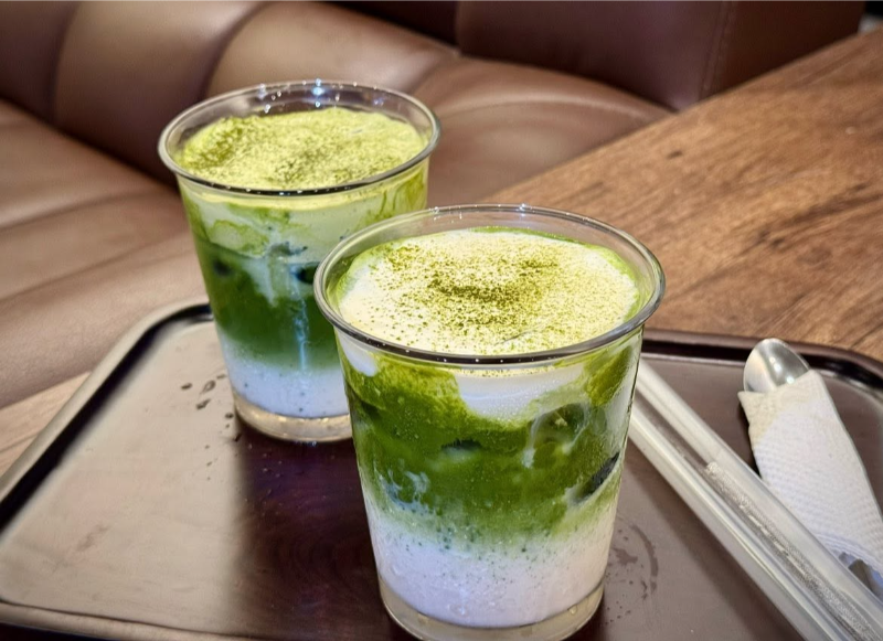 Hai ly Matcha Latte tầng lớp xanh mướt phủ lớp kem béo mịn và bột trà xanh trên khay gỗ