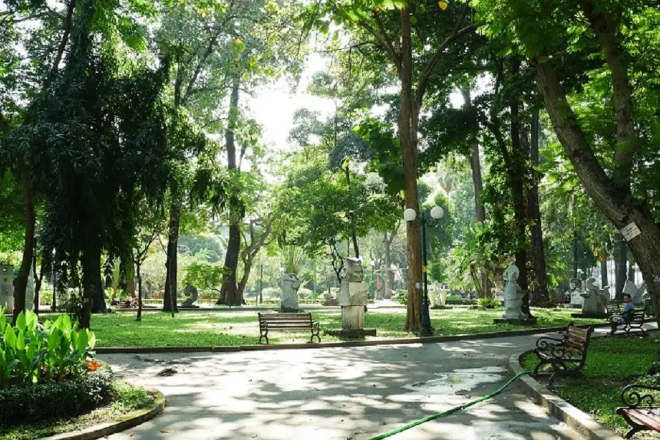 Một góc công viên với nhiều cây cổ thụ tán lá sum suê, thảm cỏ xanh mướt, lối đi bộ lát gạch, ghế đá và các bức tượng điêu khắc nghệ thuật
