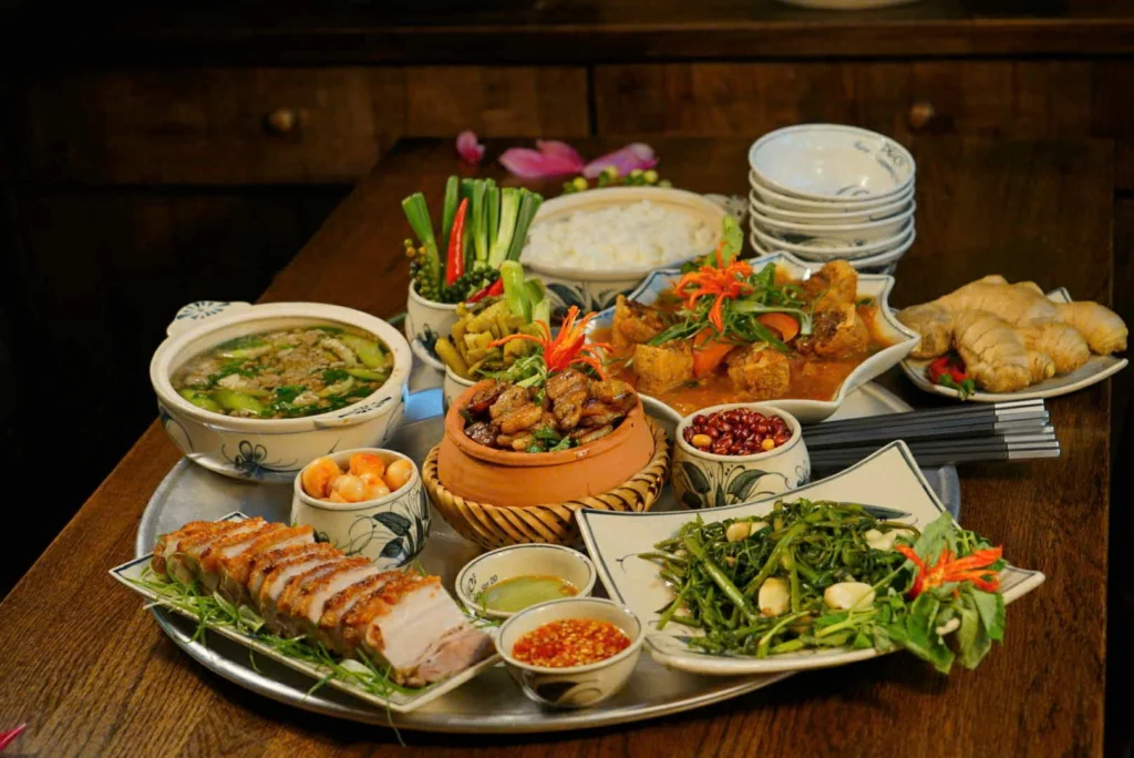Mâm cơm gia đình truyền thống Việt Nam tại một quán ăn ngon phường Đông Hưng Thuận, gồm nhiều món ăn hấp dẫn như thịt kho tộ, rau muống xào tỏi, thịt ba chỉ quay giòn, canh rau, dưa cà, lạc rang, ăn kèm cơm trắng, được bày trí đẹp mắt trên mẹt tre lót giấy bạc, xung quanh có bát đĩa cổ và gừng tươi