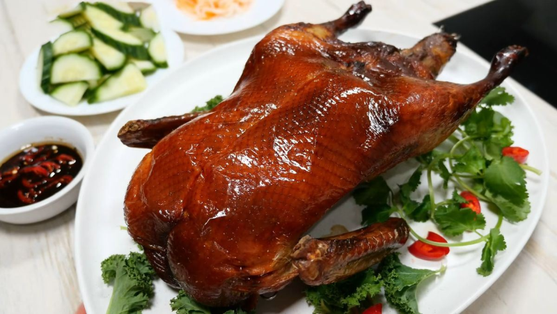 Vịt quay nguyên con da giòn rụm, bóng bẩy trên đĩa trắng, trang trí kèm rau thơm và ớt đỏ. Phía sau có đĩa dưa chuột thái lát và bát nước chấm xì dầu ớt