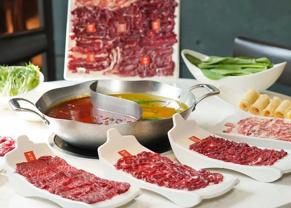 Bàn tiệc lẩu bò tươi với nồi lẩu hai ngăn, các đĩa thịt bò thái mỏng đặt trên khay hình chiếc thuyền và rổ rau xanh