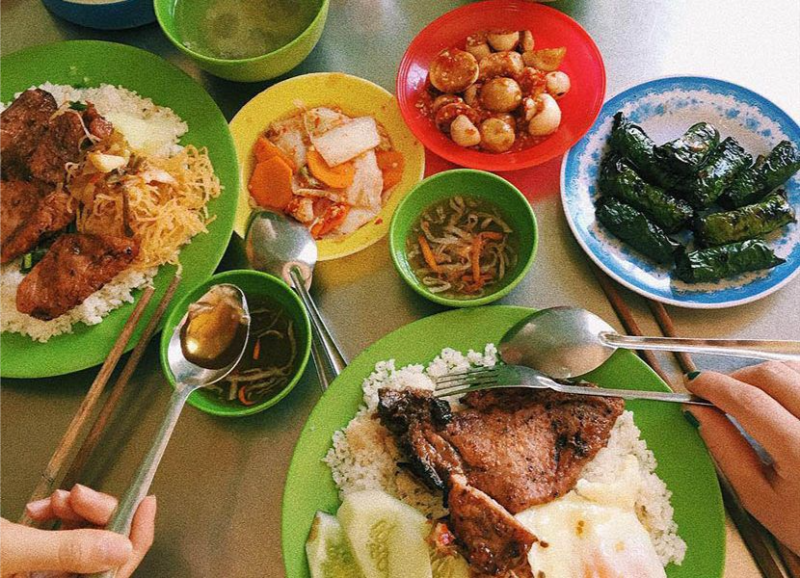 Một bữa cơm tấm Sài Gòn đầy đủ với sườn nướng, chả, trứng ốp la, dưa leo và các bát đồ chua, canh trên bàn nhôm