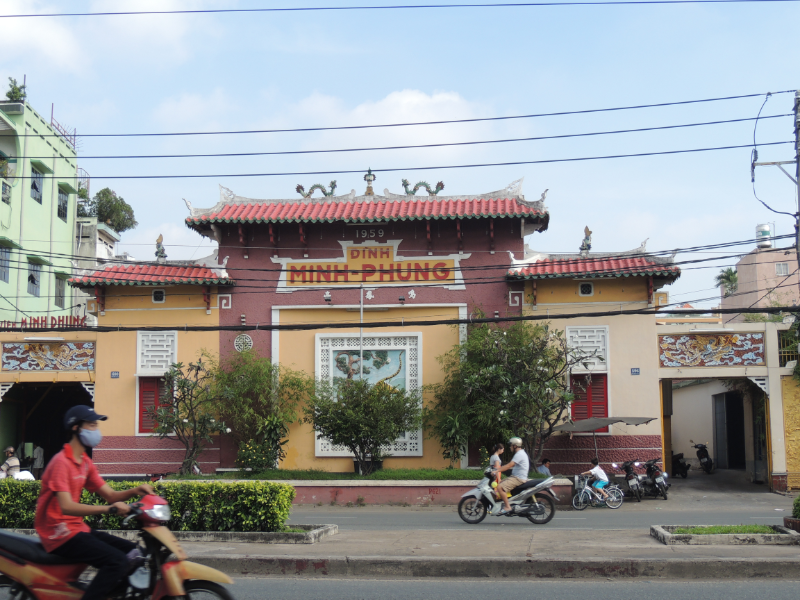 Mặt tiền Đình Minh Phụng với kiến trúc cổ kính, mái ngói đỏ, tường vàng, phía trên mái có lưỡng long chầu nguyệt. Phía trước có đường xe chạy và người đi xe máy.