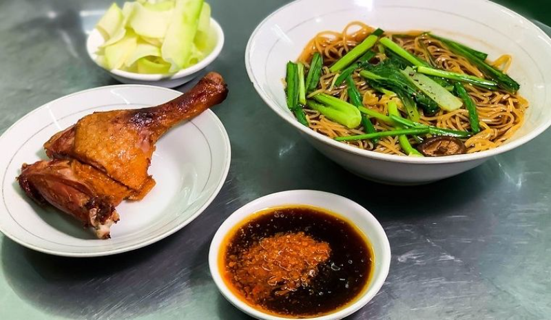 Một bát mỳ trộn vịt quay đầy đặn với sợi mỳ vàng, rau cải xanh và nấm hương. Bên cạnh là đĩa đùi vịt quay và bát nước chấm sa tế cay nồng