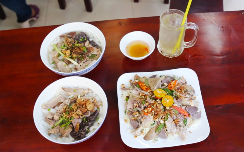 Bàn ăn gỗ bày biện các món ăn hấp dẫn: hai tô bánh canh vịt nóng hổi, một đĩa gỏi vịt trộn rau răm, hành tây, đậu phộng và một ly nước mía đá lạnh