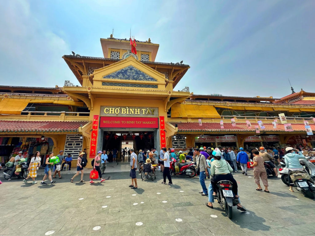 Khung cảnh nhộn nhịp của người dân và tiểu thương mua sắm trước cổng chính có kiến trúc cổ kính màu vàng đặc trưng của Chợ Bình Tây tại Quận 6 vào ban ngày