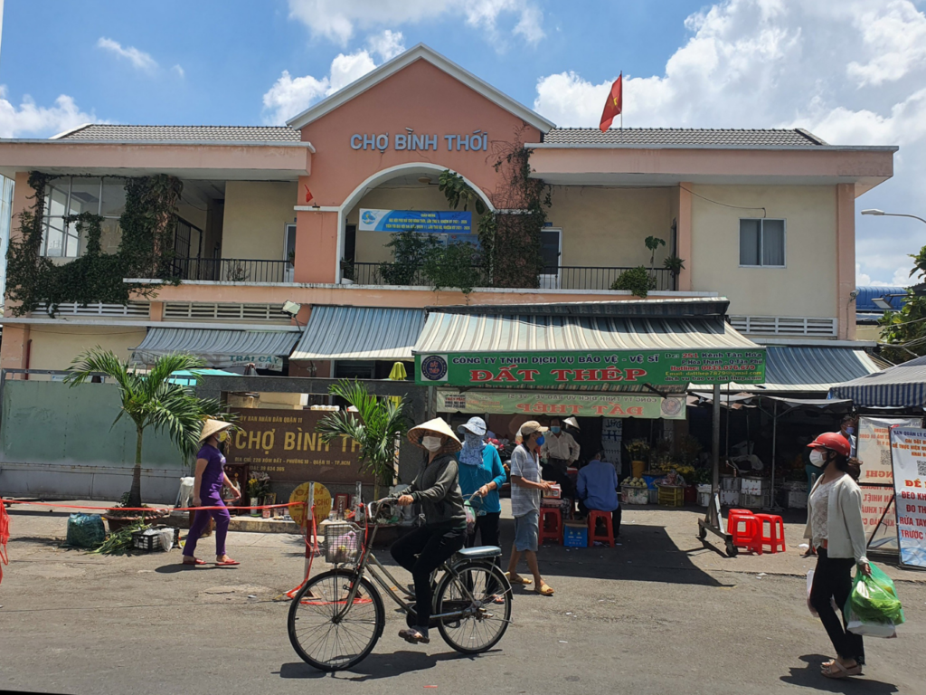 Hoạt động mua bán nhộn nhịp ban ngày trước cổng Chợ Bình Thới tại Quận 11 với người dân đi xe đạp, xe máy và các sạp hàng nhu yếu phẩm