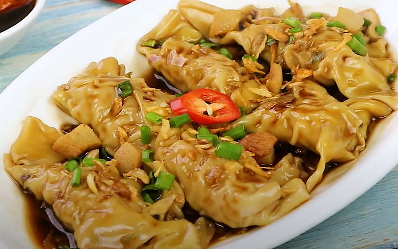 Đĩa hoành thánh trộn đầy đặn trên nền gỗ xanh. Hoành thánh được trộn với nước sốt đậm đà, rắc hành lá và hành phi, trang trí bằng một lát ớt đỏ ở giữa