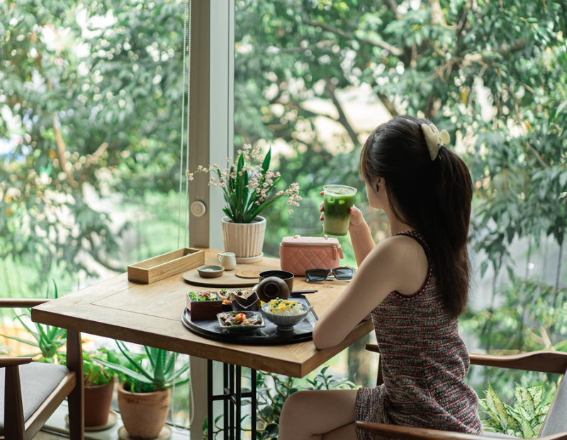 Cô gái ngồi thưởng thức ly Matcha Latte bên khung cửa sổ ngợp bóng cây xanh
