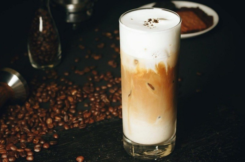 Ly thủy tinh cao đựng cà phê Bạc xỉu phân tầng rõ rệt giữa lớp sữa trắng và cà phê nâu trên nền hạt cà phê rang