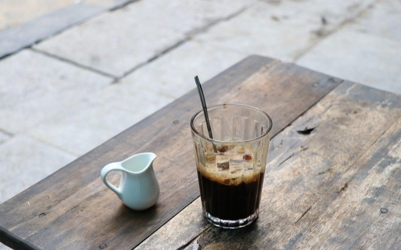 Ly cà phê đen đá pha phin sủi bọt nhẹ đặt cạnh bình rót sữa bằng sứ trên bàn gỗ mộc tại quán cafe phường Hoà Bình
